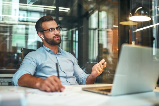 Young and successful bearded man in eyeglasses and headphones talking with client by phone while sitting in the modern office